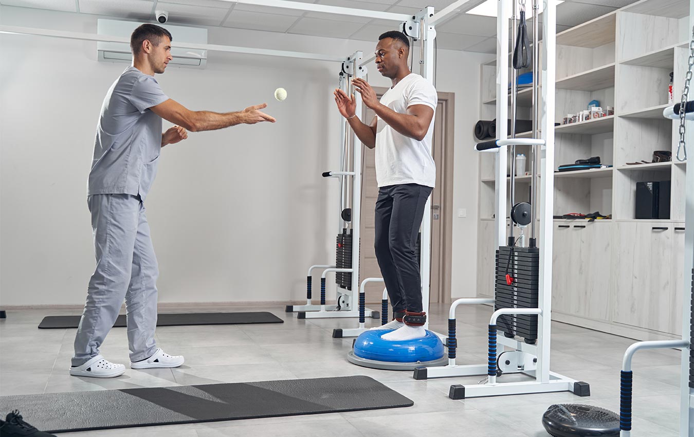 Man with ankle weights working out on balance trainer about to catch a small ball his therapist is throwing to him.