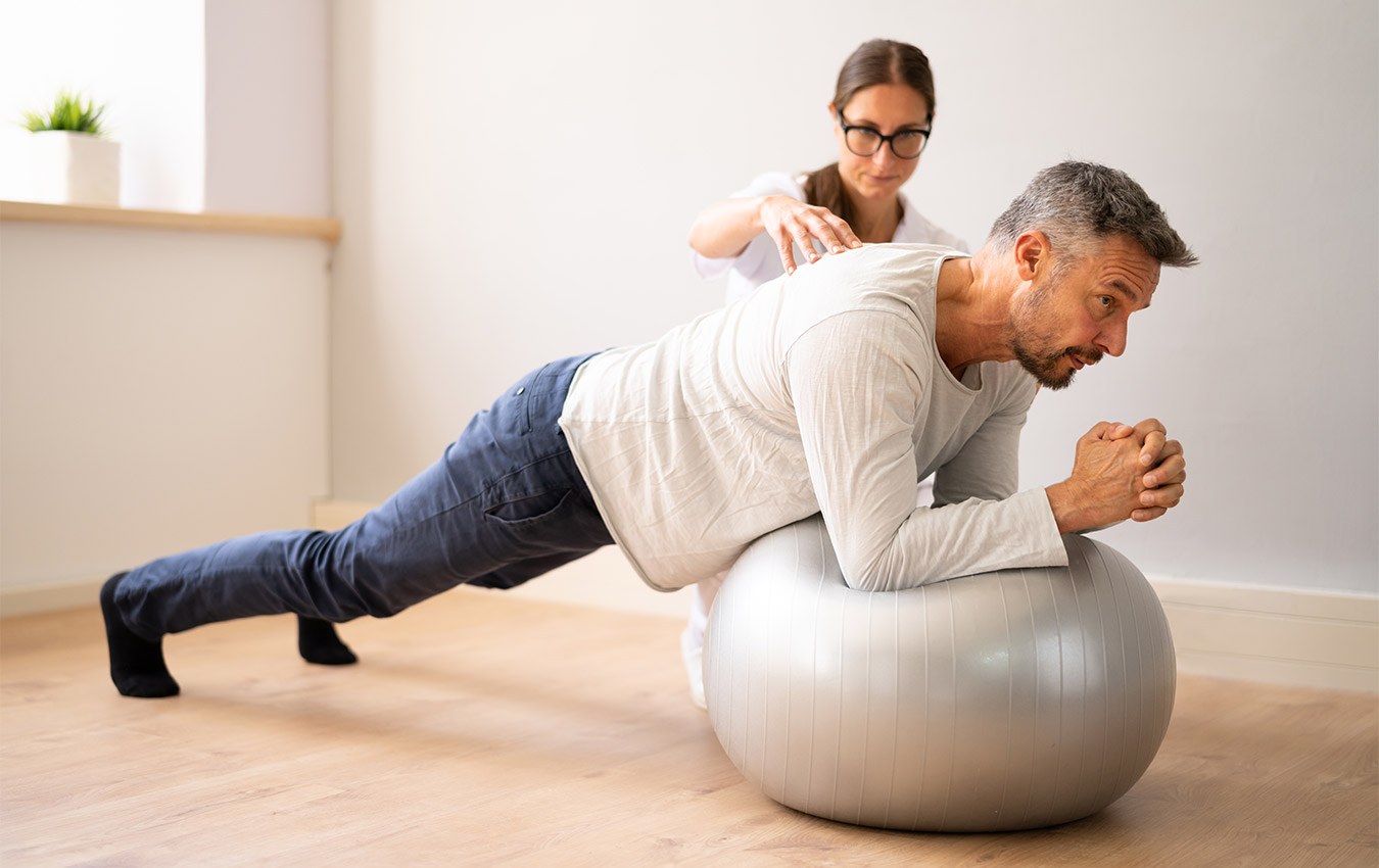 Occupational therapist correcting patient's form, while patient is doing a plank with his elbows on a medicine ball